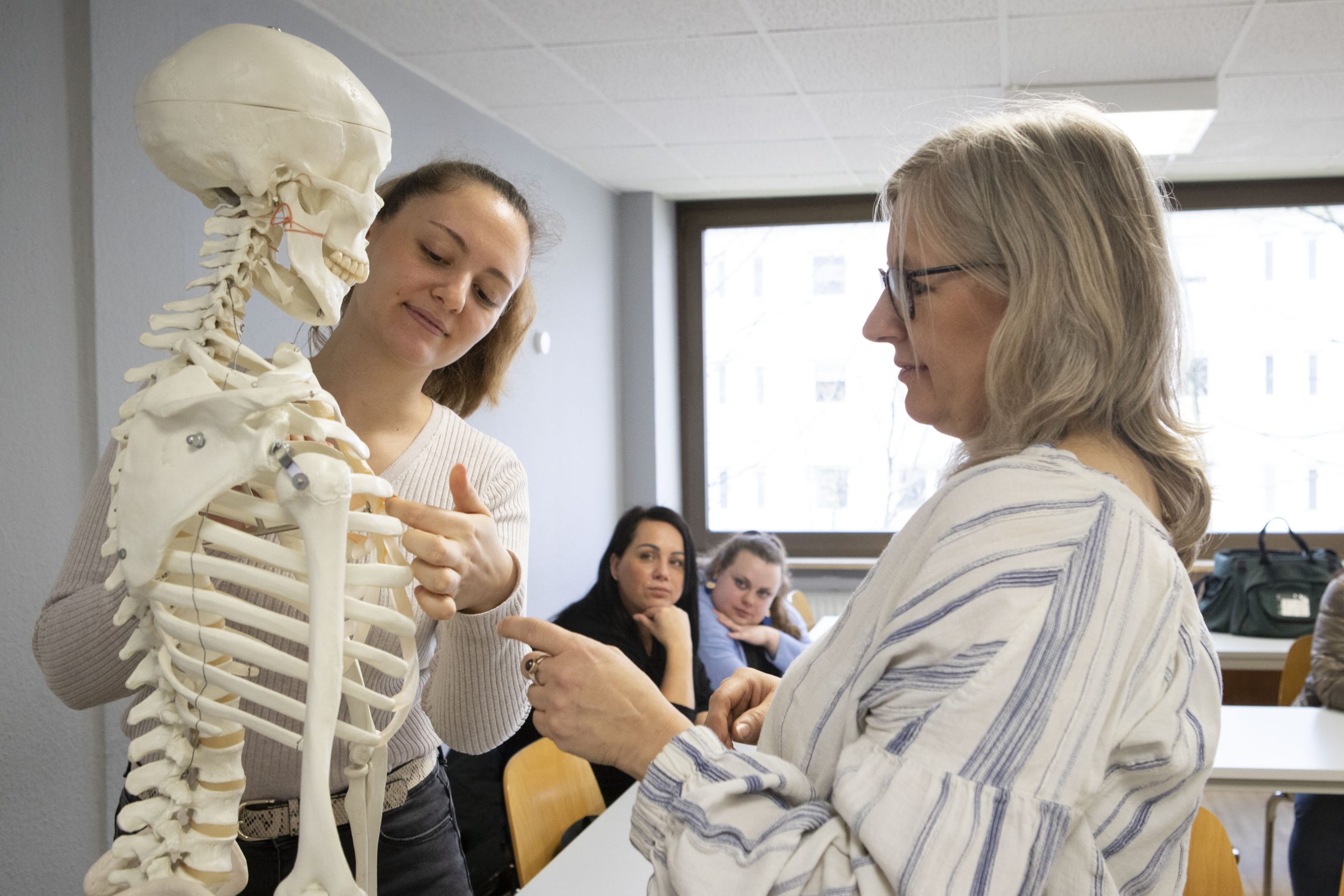 01D Lehrer und Schüler stehen in einem Klassenzimmer und begutachten ein Skelettmodell. Im Hintergrund folgen zwei weitere Schüler.