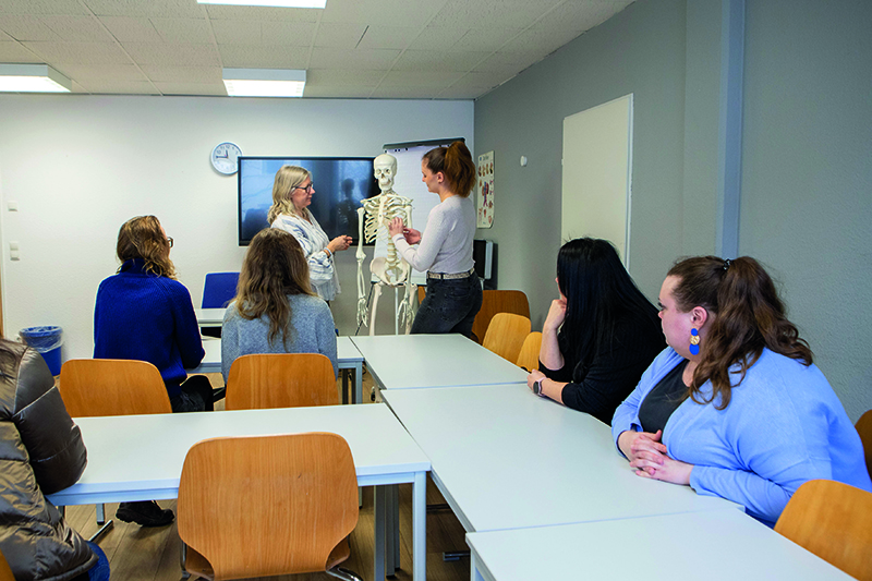 05B Der Lehrer unterrichtet die Schüler anhand des Skelettmodells. Mehrere Schüler sitzen an ihren Schreibtischen und machen mit.